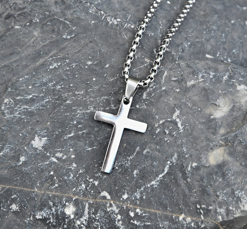 Pu&ograve; includere: Collana con pendente a croce color argento. La croce ha un design semplice e lucido. La catena &egrave; a maglie. La collana &egrave; esposta su uno sfondo grigio scuro strutturato. Simbolo religioso.