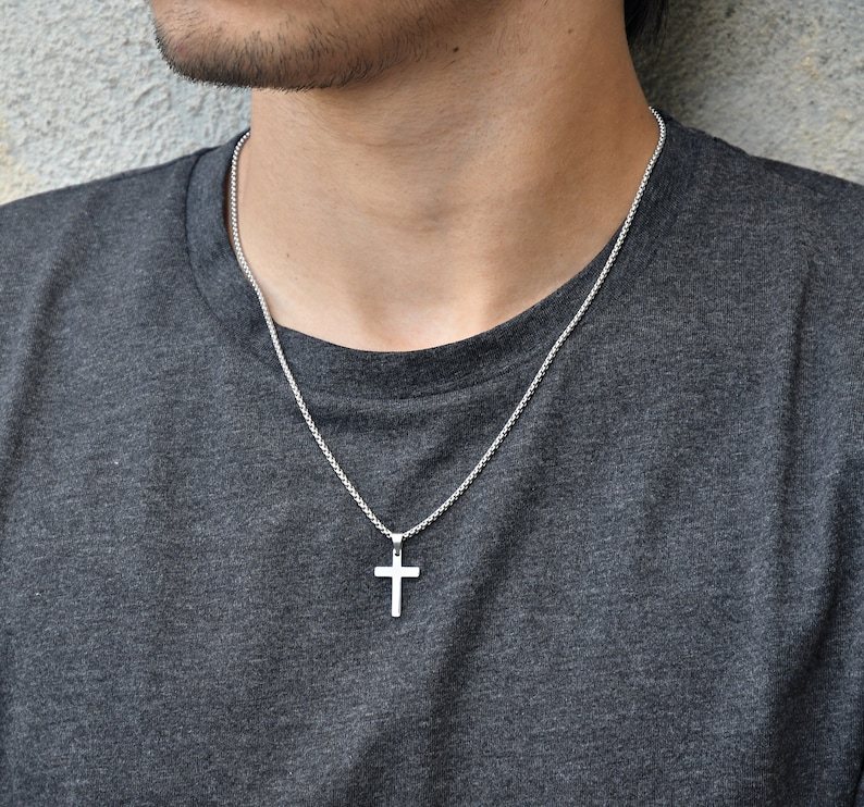 Pu&ograve; includere: Una collana con pendente a croce d'argento. La croce &egrave; un design semplice e lucido, sospeso a una catena delicata. La collana &egrave; indossata su una maglietta grigia.