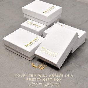May include: Several white gift boxes with gold lettering are stacked on a gray surface. The boxes have the text "Potionumber9" and a website address printed on them. A gold necklace is partially visible. The text "YOUR ITEM WILL ARRIVE IN A PRETTY GIFT BOX Ready for Gift Giving" is at the bottom.