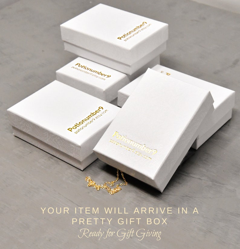 May include: Several white gift boxes with gold lettering are stacked on a gray surface. The boxes have the text "Potionumber9" and a website address printed on them. A gold necklace is partially visible. The text "YOUR ITEM WILL ARRIVE IN A PRETTY GIFT BOX Ready for Gift Giving" is at the bottom.