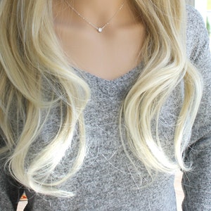 May include: A delicate silver necklace with a single, round, clear gemstone pendant. The necklace is displayed on a person wearing a gray V-neck sweater, with long, wavy blonde hair framing the neck and shoulders.