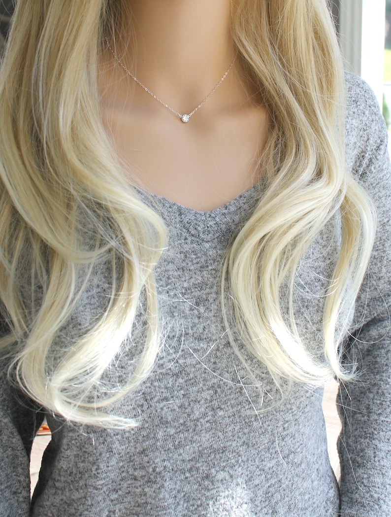 May include: A delicate silver necklace with a single, round, clear gemstone pendant. The necklace is displayed on a person wearing a gray V-neck sweater, with long, wavy blonde hair framing the neck and shoulders.