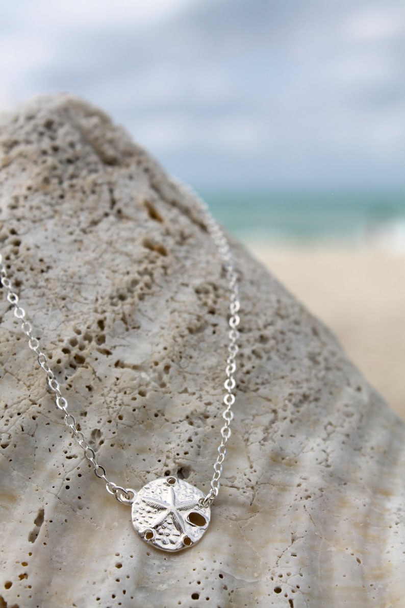 May include: A silver necklace featuring a sand dollar pendant. The pendant has a textured surface with a star design and several small holes. The necklace is displayed on a light-colored, textured rock, with a blurred beach and ocean background.