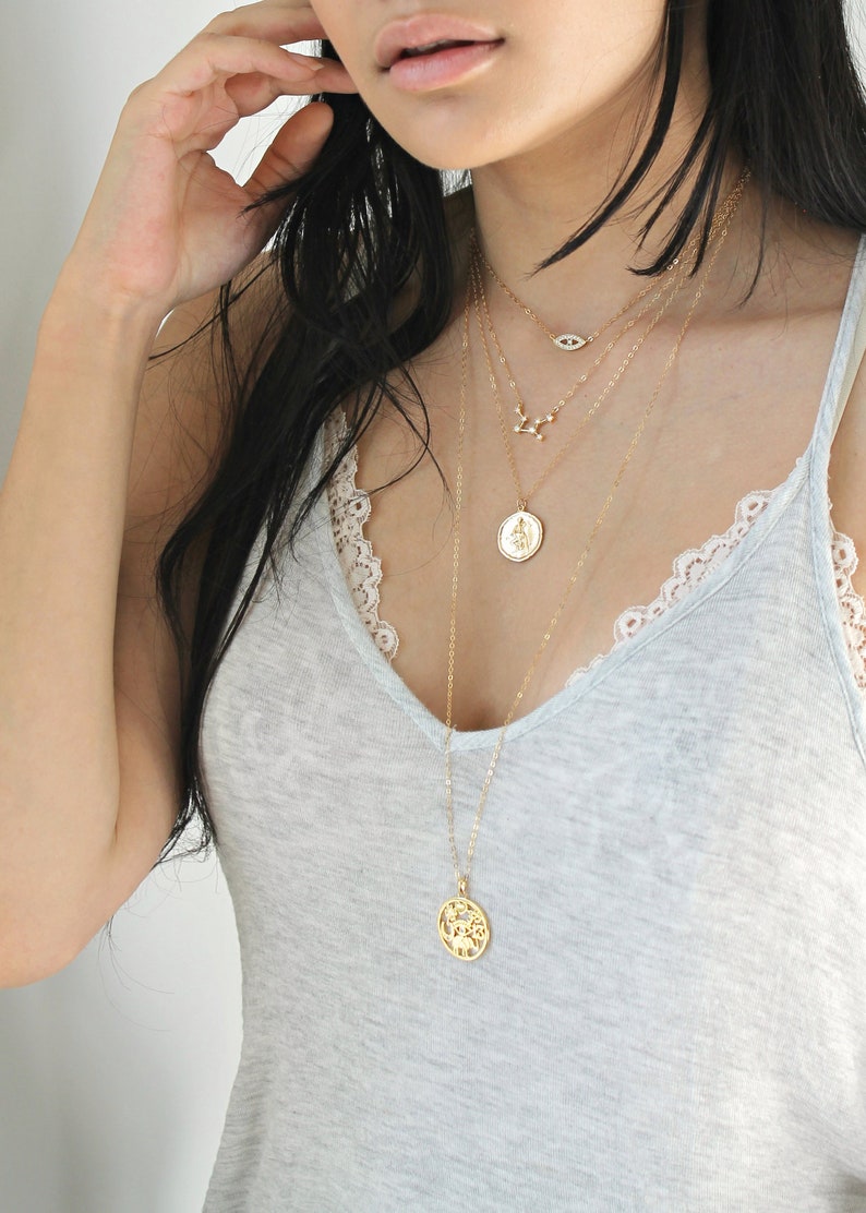 May include: A layered gold necklace set featuring four delicate chains. The necklaces include an evil eye pendant, a constellation design, a coin pendant, and a round pendant with a tree of life design. The model is wearing a gray tank top.