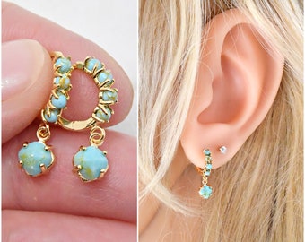 May include: Gold-tone hoop earrings with turquoise-colored stone accents. The earrings feature a circular design with small stones and a dangling, four-petal shaped stone. One earring is shown on a person's ear.
