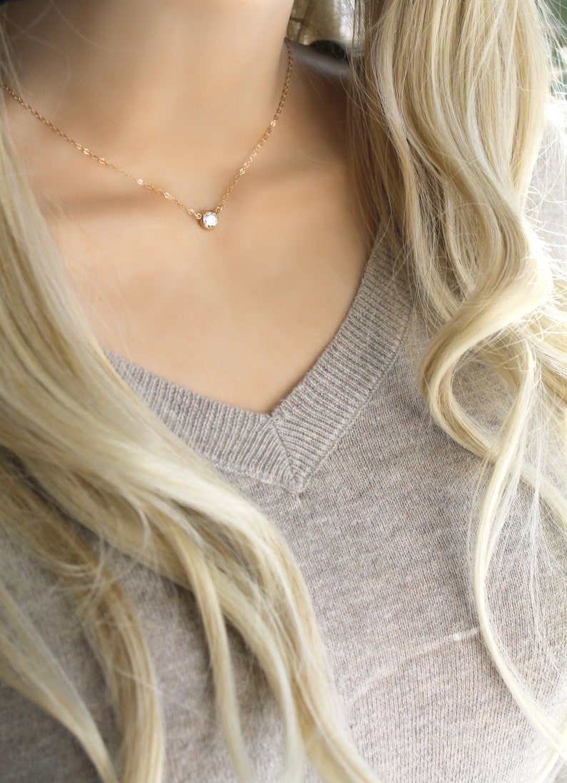 May include: A delicate gold necklace with a small, round, clear gemstone pendant. The necklace is worn against a neutral-toned v-neck sweater, with long blonde hair framing the neckline. The jewelry is simple and elegant.