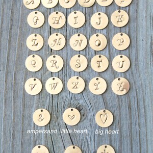 May include: Gold-colored metal charms with stylish italic uppercase letters A through Z, ampersand, hearts, dog bone, arrow, and cross. The charms are 9mm in diameter and are displayed on a wooden surface. The text "Potion No. 9" and "Stylish Italic Uppercase font" are also visible.