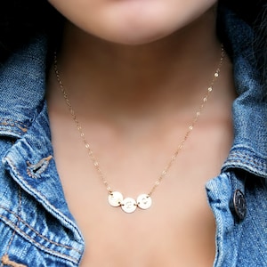 May include: A gold-colored necklace with three small, round pendants. Each pendant has a single initial engraved on it. The necklace is worn over a denim jacket.