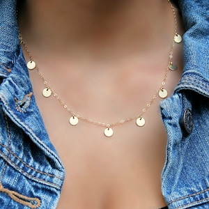 May include: A fine gold necklace with small, circular charms suspended along its length. The necklace is set against a blue denim jacket and a black top. The necklace's design is simple and elegant, suitable for everyday wear.