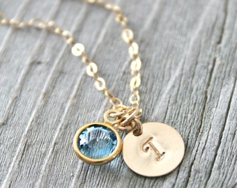 May include: Gold-toned necklace with a light blue gemstone pendant and a small, round charm with the letter "T". The necklace is displayed on a gray wooden surface. The gemstone pendant is round and set in a gold bezel.