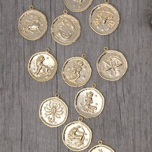 May include: Gold-colored zodiac pendants with various astrological symbols. Each pendant is round with a textured border and a small loop for a chain. The pendants are displayed on a weathered wooden surface.