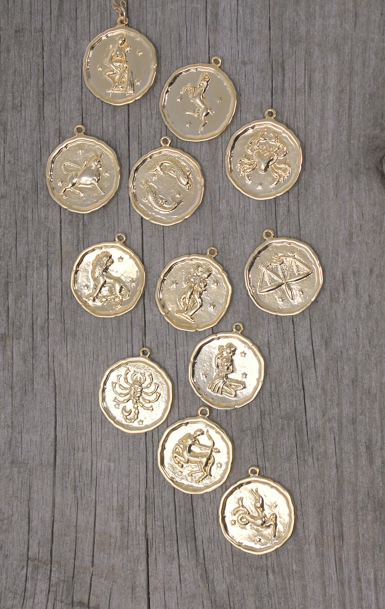 May include: Gold-colored zodiac pendants with various astrological symbols. Each pendant is round with a textured border and a small loop for a chain. The pendants are displayed on a weathered wooden surface.