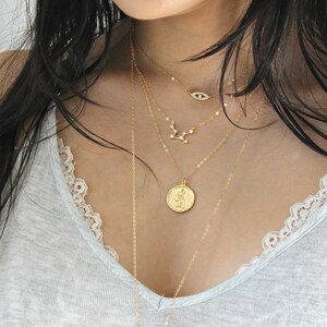 May include: A collection of layered gold necklaces. The necklaces feature various pendants, including an eye, a constellation, and a coin. The chains are delicate and the pendants are detailed. The jewelry is displayed against a light gray top.