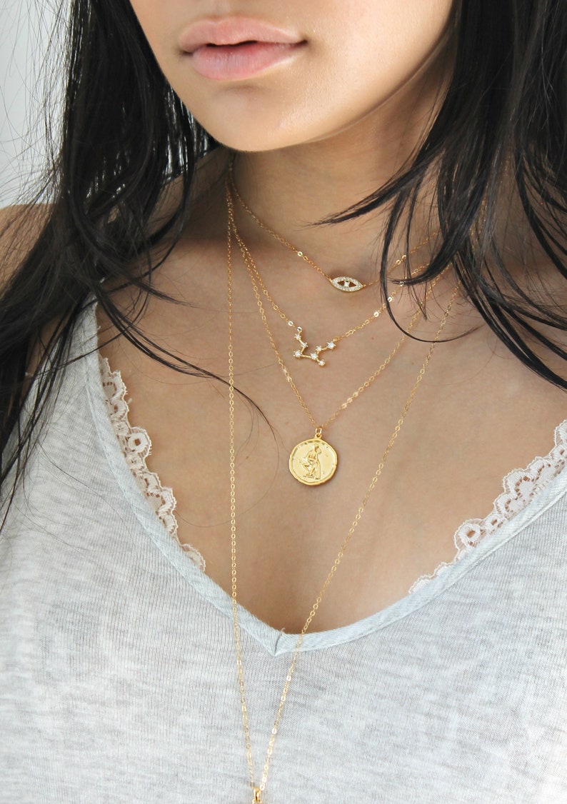 May include: A collection of layered gold necklaces. The necklaces feature various pendants, including an eye, a constellation, and a coin. The chains are delicate and the pendants are detailed. The jewelry is displayed against a light gray top.