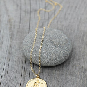May include: A gold-colored necklace with a circular pendant featuring a detailed relief design. The pendant hangs from a delicate chain, resting on a gray stone against a wooden background. The necklace is a piece of jewelry.