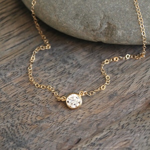 May include: A delicate gold necklace featuring a single, round, clear gemstone pendant. The pendant is set in a gold bezel and hangs from a fine gold chain. The necklace is displayed on a wooden surface.