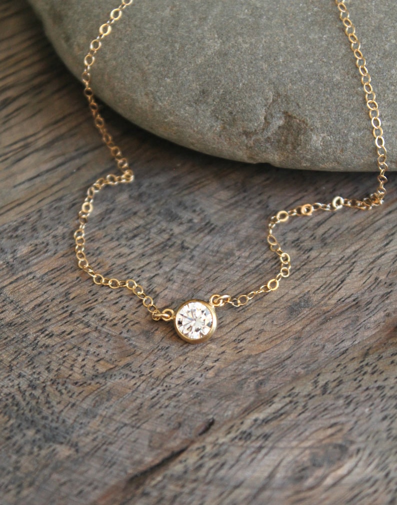 May include: A delicate gold necklace featuring a single, round, clear gemstone pendant. The pendant is set in a gold bezel and hangs from a fine gold chain. The necklace is displayed on a wooden surface.
