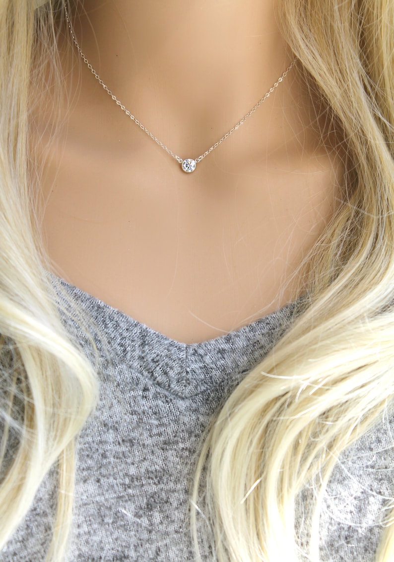 May include: A delicate silver necklace featuring a single, round, clear gemstone pendant. The pendant is suspended from a thin silver chain. The necklace is displayed on a person wearing a gray v-neck shirt.