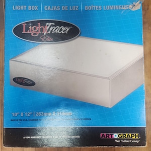 May include: A LightTracer Elite light box in a blue cardboard box with a grid pattern. The packaging displays the product name, the Artograph logo, and the dimensions 10 inches by 12 inches. The box features text in English, French, and Spanish.