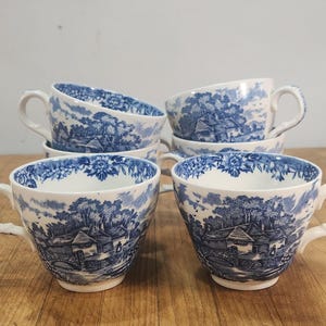 May include: A set of four white ceramic teacups featuring a blue transferware pattern. Each cup displays a detailed landscape scene with houses, trees, and a stone bridge, complemented by a floral border around the inner and outer rim.