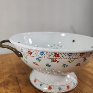 Puede incluir: Un colador de esmalte blanco con un patrón delicado de pequeñas flores rojas y azules con hojas verdes sobre un fondo punteado. El cuenco tiene dos asas de metal curvadas en los lados y una base de pedestal, con orificios de drenaje.