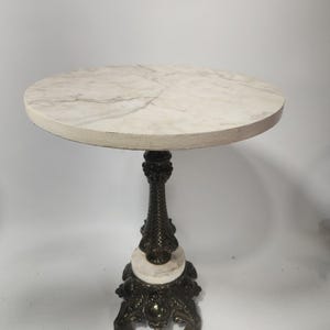 Puede incluir: Una mesa auxiliar redonda con un tablero de mármol sintético blanco con vetas grises. La base central de pedestal está hecha de metal en tono bronce oscuro con detalles intrincados, apoyada sobre una base ancha y ornamentada con un acento de mármol.