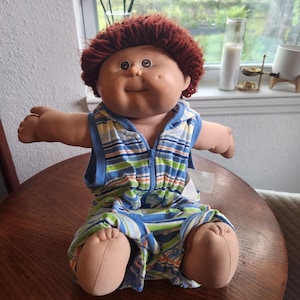 May include: A soft-bodied doll with a round face, dimpled cheeks, and short, textured reddish-brown yarn hair. The doll wears a sleeveless, hooded romper featuring a blue, green, and white horizontal striped pattern with a central blue zip.