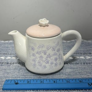 Op de afbeelding: Een crèmekleurige keramische theepot met een lichtroze deksel met een kleine bloemvormige knop. Het lichaam is versierd met een herhalend patroon van kleine paarse bloemen en heeft een zichtbare craquelé glazuurlaag. Een blauwe liniaal toont 15 cm.