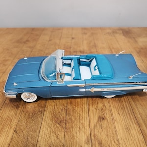 Könnte beinhalten: Ein blaues Modellauto als Cabriolet mit weiß-blauem Innenraum. Das Fahrzeug verfügt über eine klare Windschutzscheibe, silberfarbene Zierelemente, Weißwandreifen und den Schriftzug Impala in Silber auf dem hinteren Seitenteil.