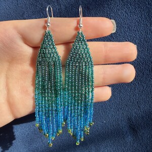 May include: A pair of long, dangling earrings crafted from small, iridescent glass beads. The beads transition in color from a deep emerald green at the top to a vibrant sky blue at the bottom, finished with small metallic gold accent beads.