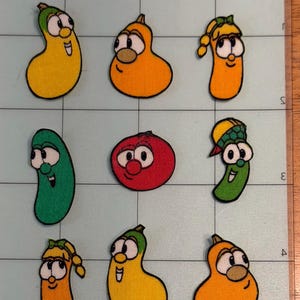 May include: A set of nine embroidered fabric patches featuring cartoon vegetable characters. The collection includes yellow, orange, green, and red shapes with black outlines, arranged in a grid on a light blue cutting mat with grid lines.