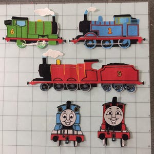 May include: A collection of five felt train cutouts featuring three side-view locomotives and two front-view faces. The green train is numbered 6, the blue train is numbered 1, and the red train is numbered 5. Each train includes white smoke puffs.