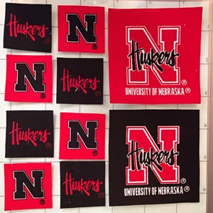 May include: A collection of square fabric patches featuring University of Nebraska branding. The patches include red and black designs with the letter N, the word Huskers in script, and the full text University of Nebraska on a cutting mat.
