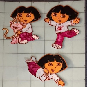 May include: Three embroidered fabric patches featuring the cartoon character Dora. One patch shows Dora hugging a small monkey, another shows her standing with arms outstretched, and the third shows her lying down. All feature black hair and pink pants.