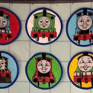 May include: Six circular fabric patches featuring cartoon train characters. The top row shows a blue train on red, a green train on white, and a blue train on light blue. The bottom row shows a train on light blue, a red train on green, and a train on yellow.