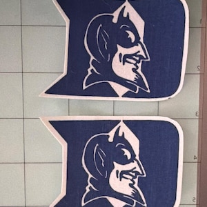 May include: Two identical blue and white vinyl decals shaped like the letter D, each featuring a profile illustration of a devil mascot with horns and a pointed chin. The decals are arranged vertically on a light green grid cutting mat.