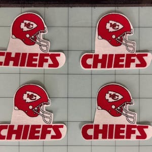 May include: Four identical embroidered patches featuring a red football helmet with a white arrowhead logo and the word CHIEFS in bold red capital letters. The patches are arranged in a grid on a cutting mat with visible grid lines and numbers.