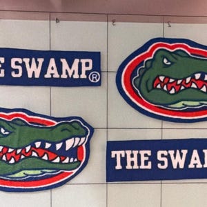 May include: Two rectangular blue patches with white text reading THE SWAMP and two oval patches featuring a green alligator head with white teeth and red accents. The patches are arranged on a white grid surface with visible black lines.
