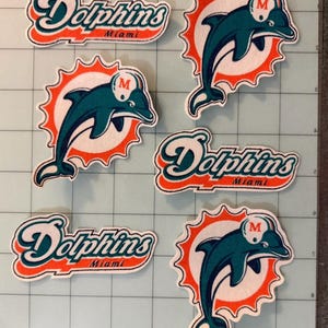 May include: Three embroidered patches featuring the Miami Dolphins logo, a teal dolphin wearing a white helmet with an orange M, set against an orange sunburst. Three additional patches display the text Dolphins Miami in teal and orange.