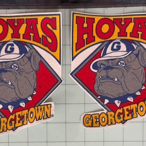 May include: Two identical adhesive decals featuring a brown bulldog wearing a blue and white baseball cap with a G logo. The design includes a red diamond shape, the word HOYAS in red letters, and the word GEORGETOWN in yellow letters on a blue background.