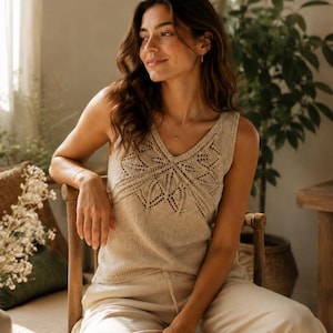 May include: A sleeveless beige knit top featuring a V-neckline and an intricate lace-like pattern across the chest. The garment is constructed with a fine-gauge knit texture and a straight hemline, providing a neutral-toned piece of clothing.