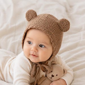 May include: A light brown knitted bonnet featuring two small rounded bear ears on top and matching chin ties. The bonnet is paired with a small, light brown knitted teddy bear toy that has a stitched nose and mouth, held by a baby.
