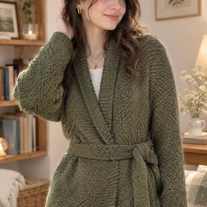 May include: An olive green knit cardigan featuring a shawl collar, long sleeves, and a matching tie belt at the waist. The garment has a textured, chunky knit pattern throughout and is worn over a plain white top with light-colored trousers.