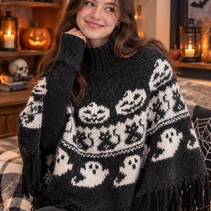 Könnte beinhalten: Ein schwarzer Strickponcho mit einem weißen Jacquardmuster aus Kürbissen, schwarzen Katzen und Geistern. Das Kleidungsstück hat einen hohen Rippkragen, lange Ärmel und einen Saum, der mit langen schwarzen Fransen verziert ist.