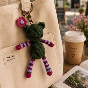 May include: A dark green crocheted frog keyring with striped purple and pink arms and legs, attached to a beige canvas tote bag. The bag features a rectangular patch with the text "merci beaucoup" stitched in black on the front pocket.