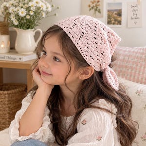 May include: A light pink triangular crochet headscarf featuring an intricate openwork diamond pattern and a knotted tie closure at the back. The accessory is crafted from yarn and designed to be worn over the hair, covering the top of the head.