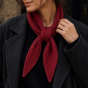 May include: A deep red knitted triangular scarf tied in a simple knot at the neck. The scarf features a textured garter stitch pattern with pointed ends. It is worn over a black sweater and a dark charcoal gray wool coat with a notched lapel.