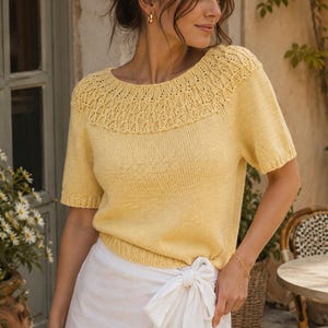 May include: A pale yellow short-sleeved knit sweater featuring a round neckline with an intricate, textured lace-like pattern across the yoke and shoulders, paired with a white wrap-style skirt that ties at the waist with a fabric bow.