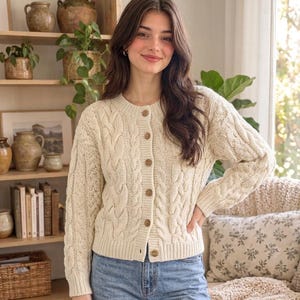 May include: A cream-colored cable knit cardigan featuring a round neckline, long sleeves with ribbed cuffs, and a front button closure with six brown buttons. The garment is paired with light blue denim jeans.