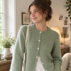 May include: A sage green knit cardigan featuring a crew neckline, long sleeves, and a front button closure with white buttons. The cardigan is worn over a plain white top and paired with white drawstring trousers, accompanied by a floral tote bag.